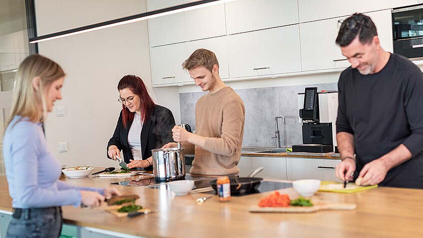 Vier Personen stehen in einer Küche an einer Arbeitsfläche und schneiden gemeinsam Gemüse etc. klein